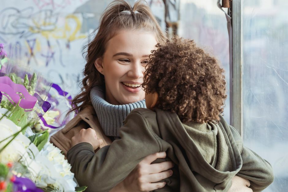 A young woman with wavy, light brown hair and a grey turtleneck sweater smiling warmly as she embraces a child with curly brown hair wearing a green jacket, surrounded by an array of fresh, colorful f