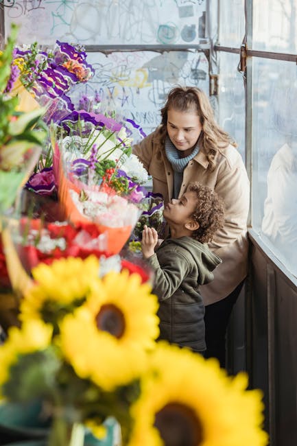 A woman and a young child examining a vibrant display of fresh flowers at a florist shop. The arrangement includes a variety of colorful blooms such as purple irises, red and pink roses, yellow sunflo