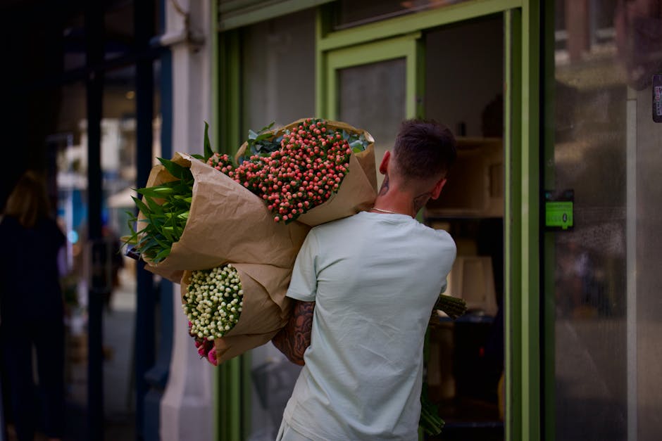 A person with short brown hair, wearing a light grey T-shirt, is carrying a large floral bouquet wrapped in brown paper, featuring clusters of pink and white berries and green foliage. The bouquet is