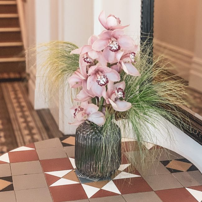 A tall floral arrangement featuring soft pink orchids with white and maroon markings on their petals, placed in a dark, ribbed glass vase. The orchids are accompanied by wispy green foliage and delica