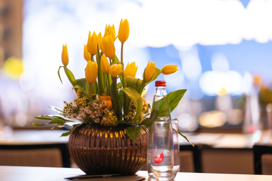 A floral arrangement featuring vibrant yellow tulips with closed and partially opened petals, arranged in a round, dark ribbed glass vase. The bouquet is complemented by smaller pink and white filler