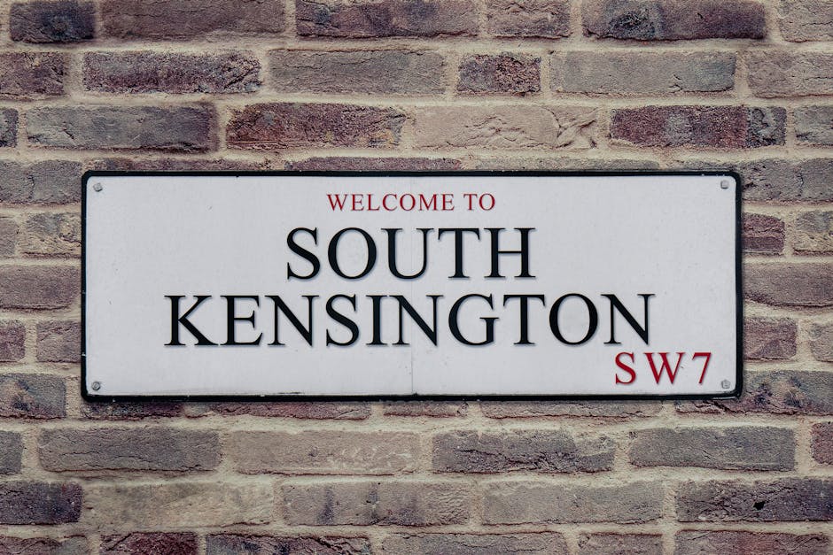 A rectangular white metal street sign mounted on a red-brown brick wall, with black text reading 'Welcome to South Kensington', and a smaller red inscription 'SW7' in the bottom right corner, set in a