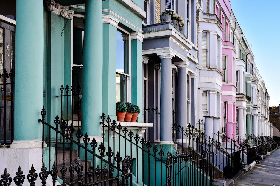 A row of colorful Victorian-style terraced houses in Ladbroke Grove near Notting Hill Gate, featuring pastel-painted facades in shades of turquoise, gray, pink, and white. The houses have large sash w