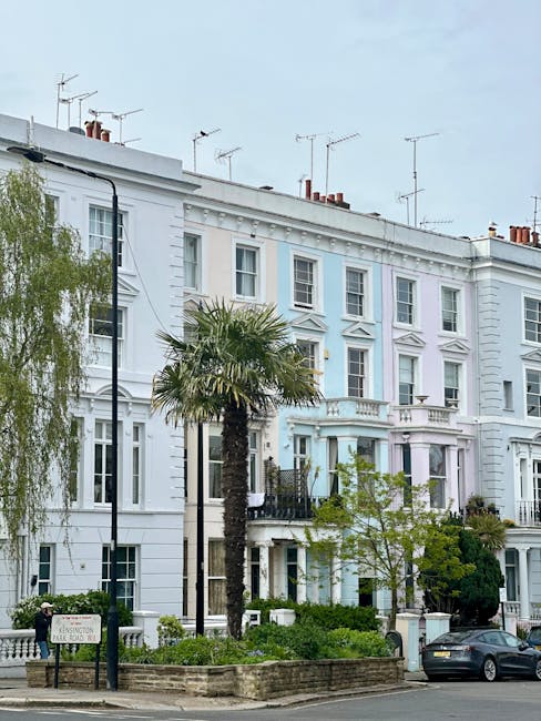 A row of elegant, white Victorian-style townhouses along a street in Notting Hill, featuring classic architectural details such as decorative cornices and bay windows. In the foreground, a landscaped