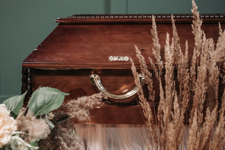 A polished wooden casket with a dark finish and silver handles, situated in a softly lit room surrounded by tall, beige pampas grass and subtle greenery. The arrangement includes cream and blush flora