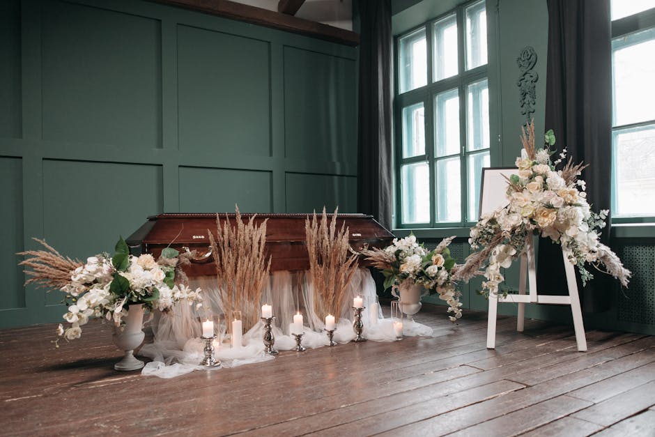 A floral arrangement featuring white roses, orchids, and assorted greenery, complemented by tall pampas grass and soft, flowing fabrics, is displayed on a wooden floor in front of a green paneled wall