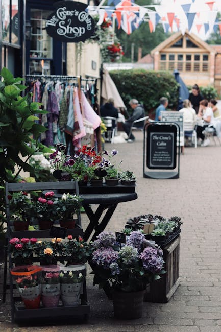 An outdoor display of potted flowering plants and small bouquets situated on black and wooden tables outside The Rose's Garden Shop, with vibrant purple and pink hydrangeas, red and white begonias, an