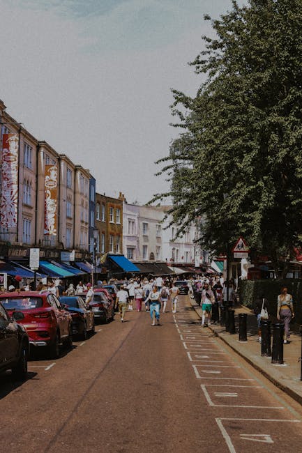 The image shows a lively street scene with pastel-colored shopfronts and a bustling crowd of pedestrians. Parked cars line both sides of the narrow road, which features white roadside markings. On the