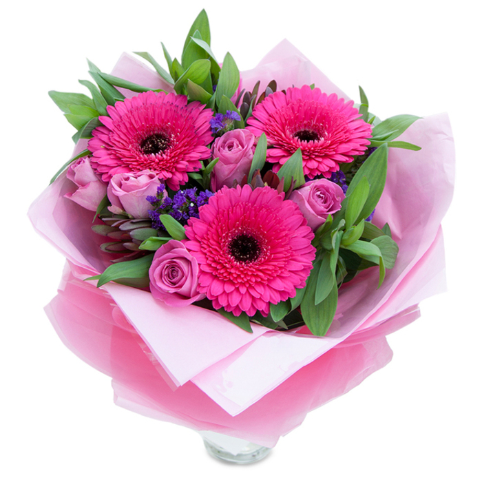 I picture this arrangement on a small kitchen table, the three glossy fuchsia Gerberas open and proud, their dark centres punctuating the hot-pink intensity, while lavender-tinged roses curl inward with the tender texture of new growth. Tiny clusters of purple statice tuck in between the focal blooms, adding playful, papery contrast and a slightly wild, meadow-like character. Surrounding greenery-broad, glossy leaves that read as salal or eucalyptus-creates a cool, verdant frame and a subtle green fragrance that balances the roses' softness. The bouquet is lovingly cocooned in a pale blush tissue layer and finished with a translucent, slightly crinkled fuchsia-pink cellophane wrap that catches the light in thin ribbons, giving a graceful shimmer to the whole piece; the folds are neat, intentionally arranged to present the flowers like a gift. At the bottom you can see the top rim of a clear glass vase, suggesting immediacy and readiness to display. The scent profile is gentle: roses lend a soft perfume, gerberas a fresh, almost green hint, and the foliage adds an elemental, clean note. As a florist familiar with the rhythms of Portobello Road market and the calm corners of Holland Park, I chose these tones and textures to convey playfulness and grace-an arrangement that suits a celebratory morning, a bedside surprise, or an anniversary table. The overall effect is bright, intimate, and thoughtfully composed to make someone feel cherished.